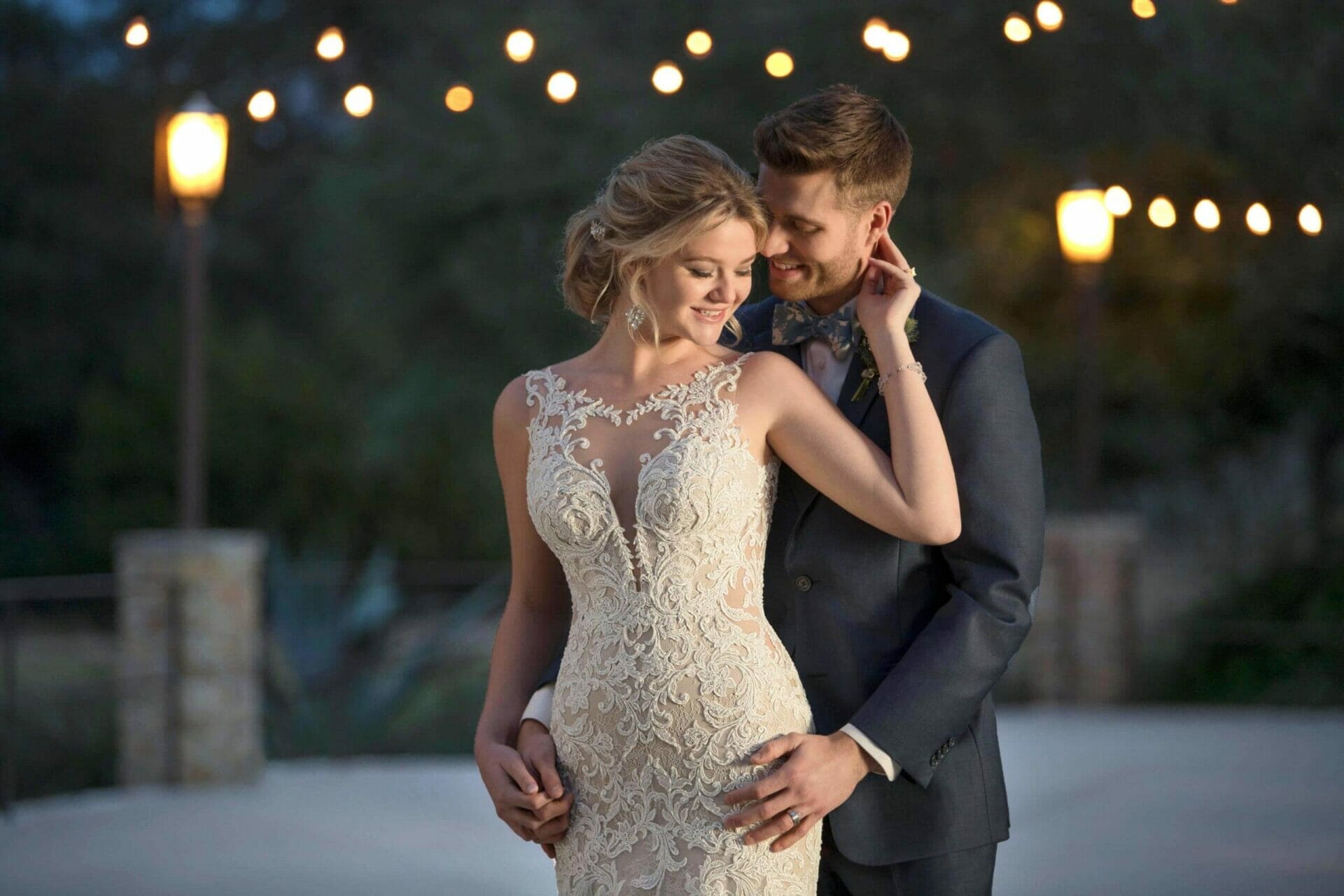 A bride and groom embracing in front of a string of lights outdoors at their wedding reception.