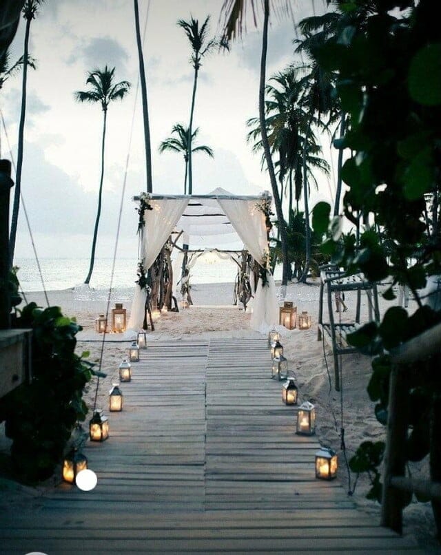 A wooden walkway leading to a beach with candles and palm trees, perfect for a romantic wedding ceremony.