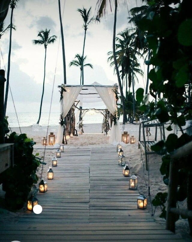 Destination Decompress A wooden walkway leading to a beach with candles and palm trees, perfect for a romantic wedding ceremony.