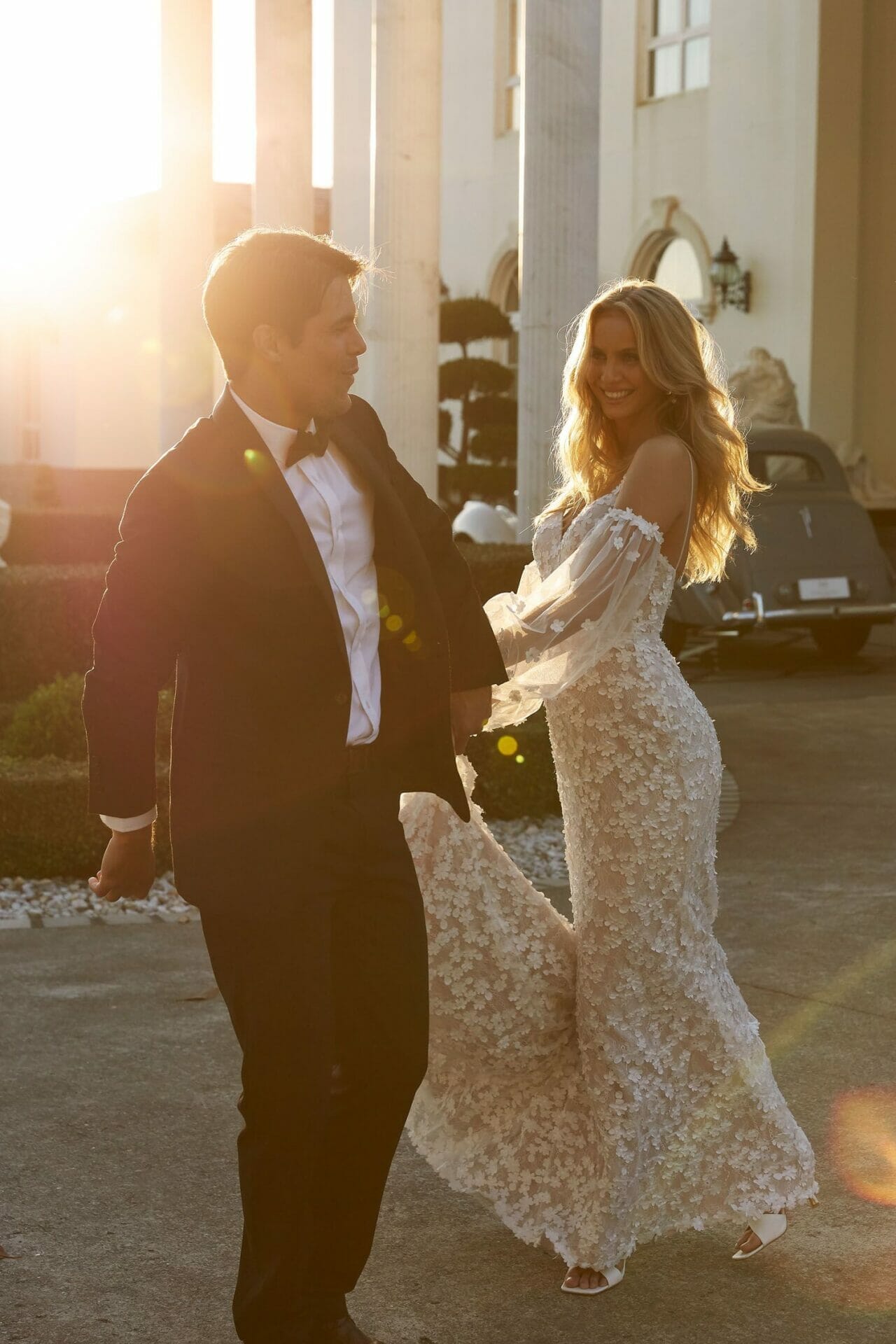 A bride and groom, dressed in wedding attire, walking in front of a building at sunset.