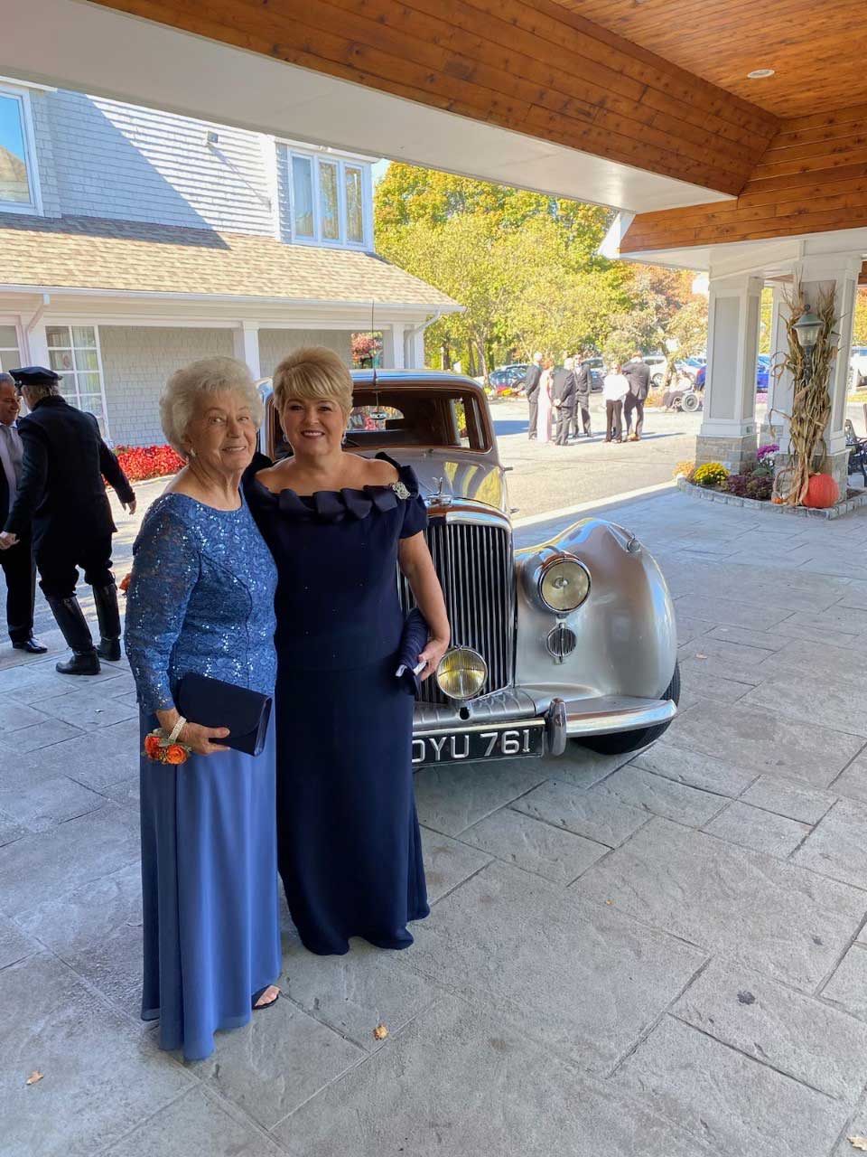 Two women standing in front of a vintage car outside a bridal store.