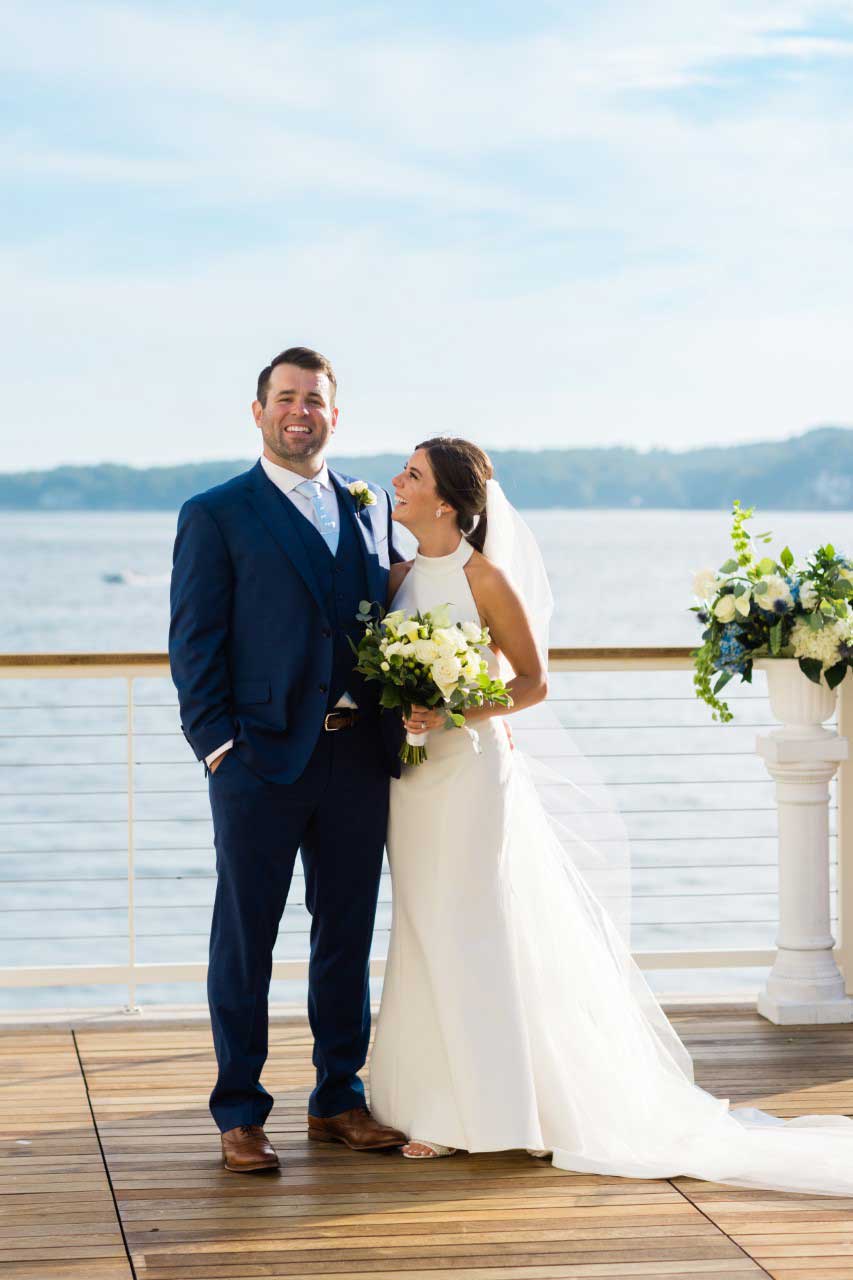 A bride and groom standing on a deck overlooking the water at their wedding.