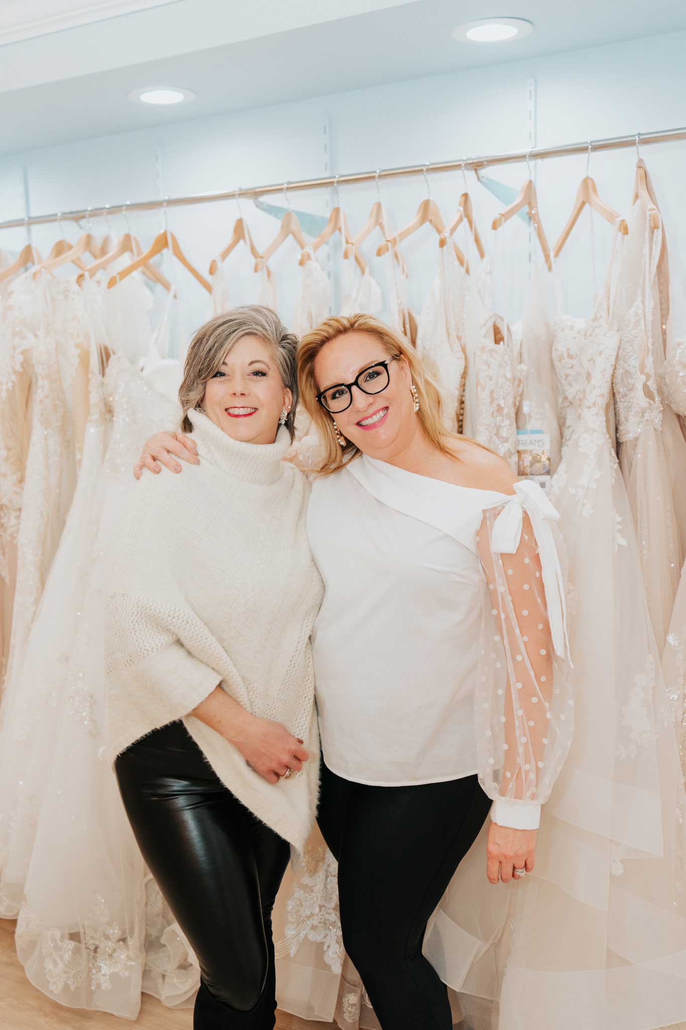 Two women posing for a photo in a bridal shop, surrounded by wedding dresses.