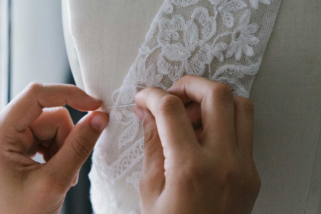 alterations-1097786566 A person is embellishing a wedding dress with lace at a bridal salon.