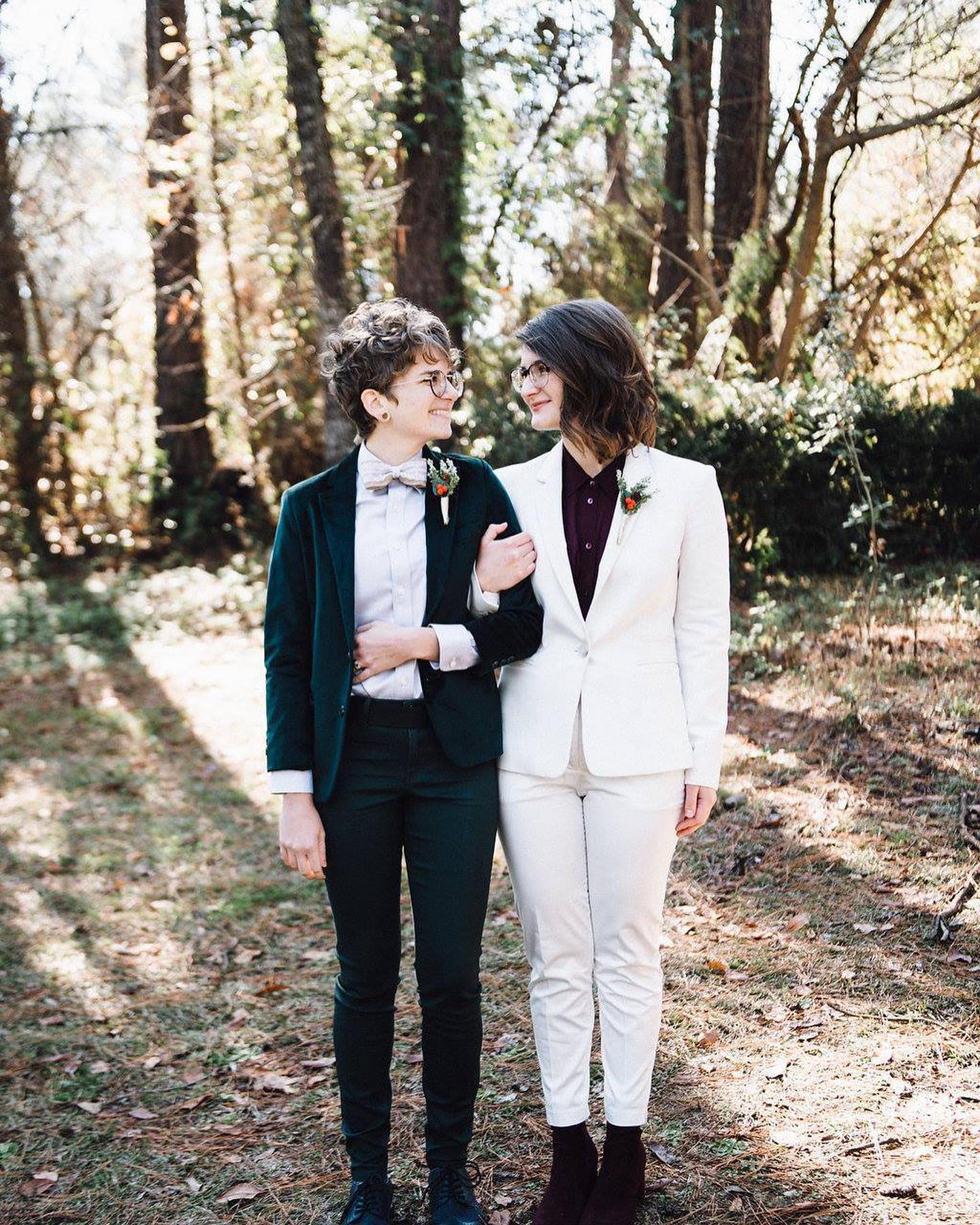 gender-inclusive-wedding-suits Two women in suits standing in a wooded area near a bridal salon.