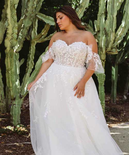 A plus size woman wearing a white wedding dress in front of cactus at a bridal salon.