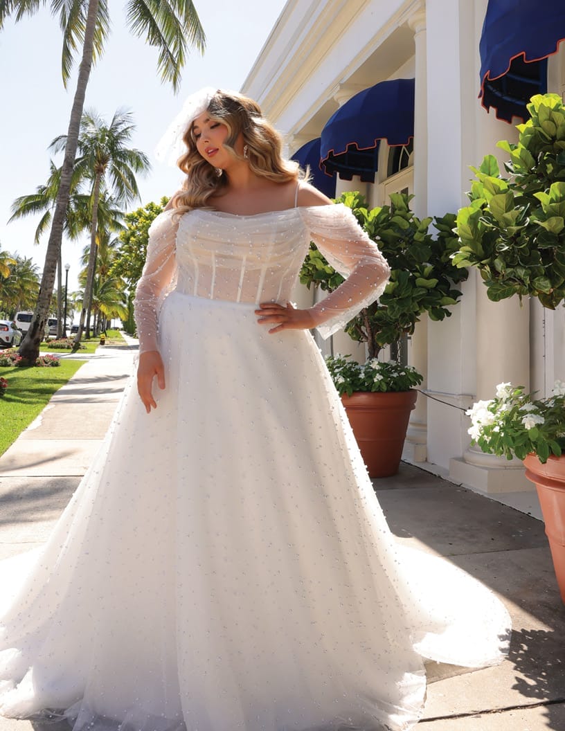 A woman wearing an elegant off-shoulder wedding dress with long sleeves, posing in a sunlit street lined with palm trees and elegant buildings.