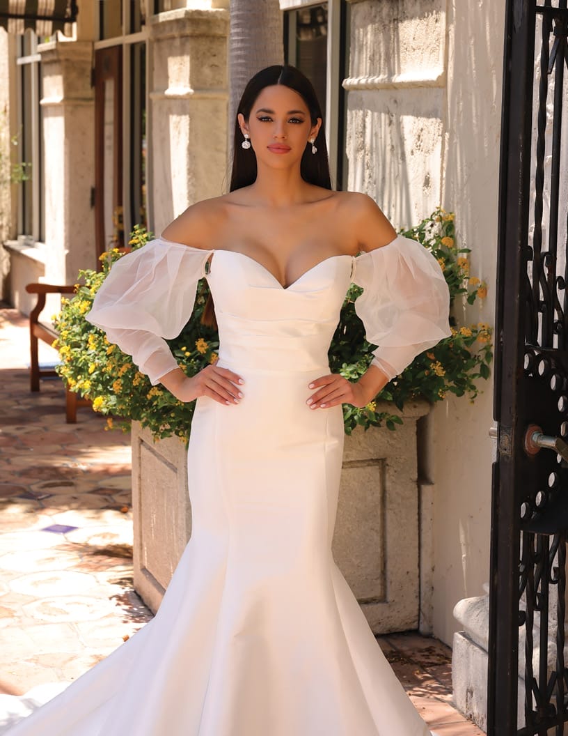 A woman in an elegant white off-the-shoulder gown with sheer sleeves standing in a sunlit courtyard.