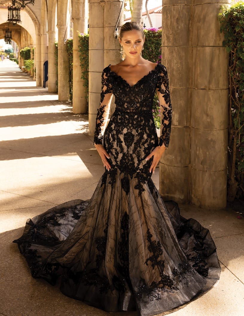 A woman in an elegant black lace gown stands confidently on a sunlit pathway with arches in the background.
