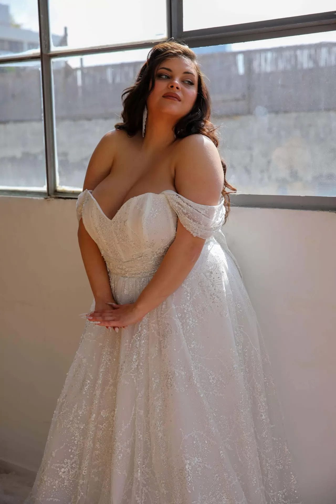 A woman in an off-shoulder white wedding dress stands indoors, leaning slightly forward with hands together. She has long, wavy hair and is looking towards the window.