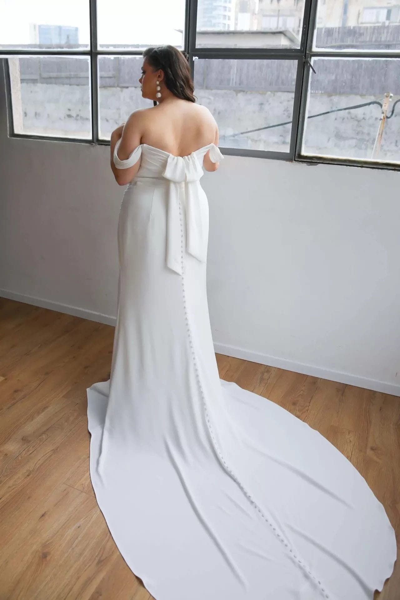 A woman in a white off-shoulder gown with a long train stands facing a window in a room with wooden floors.