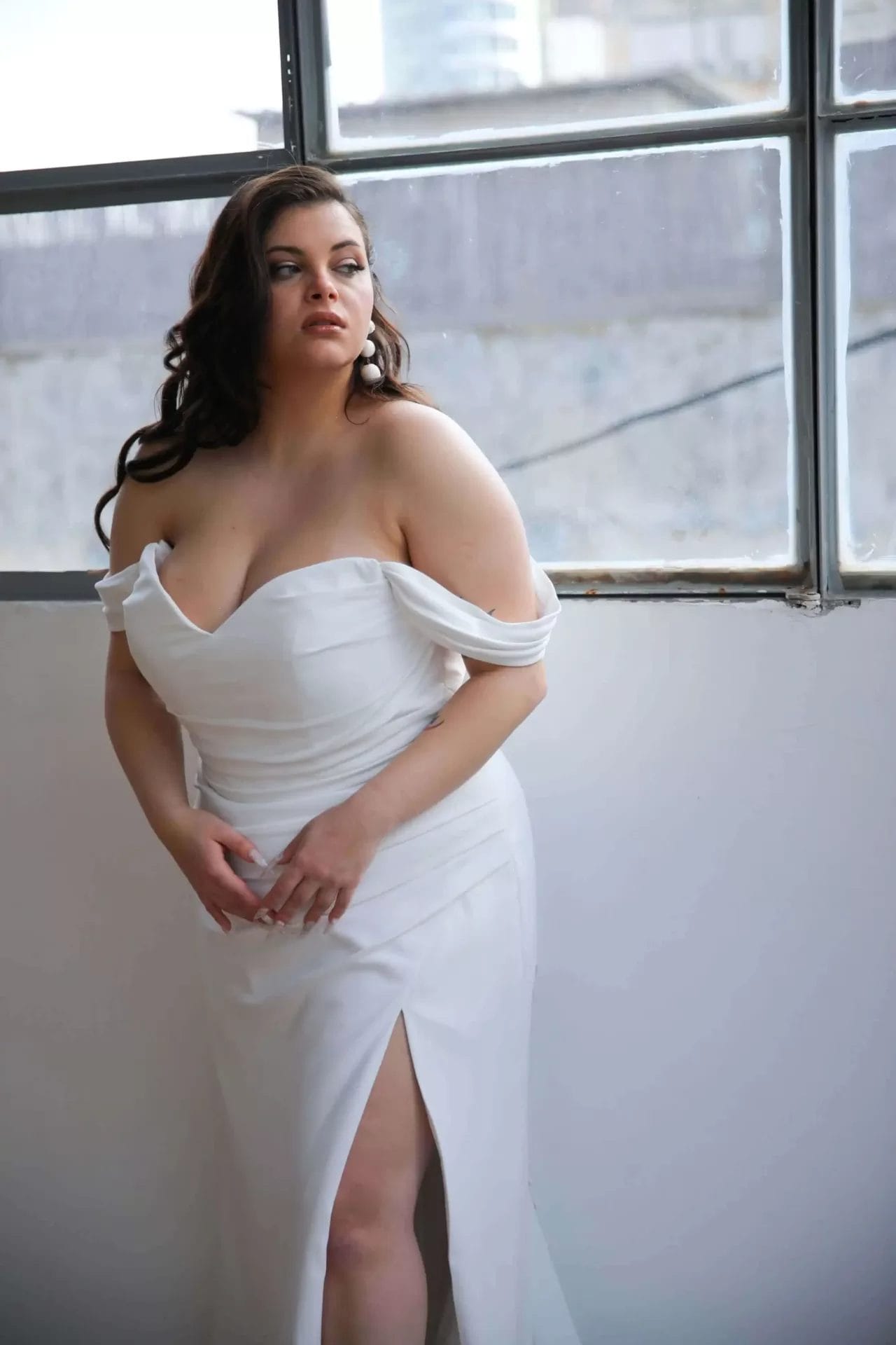Person in an off-the-shoulder white dress with a thigh-high slit stands by a window, looking to the side.