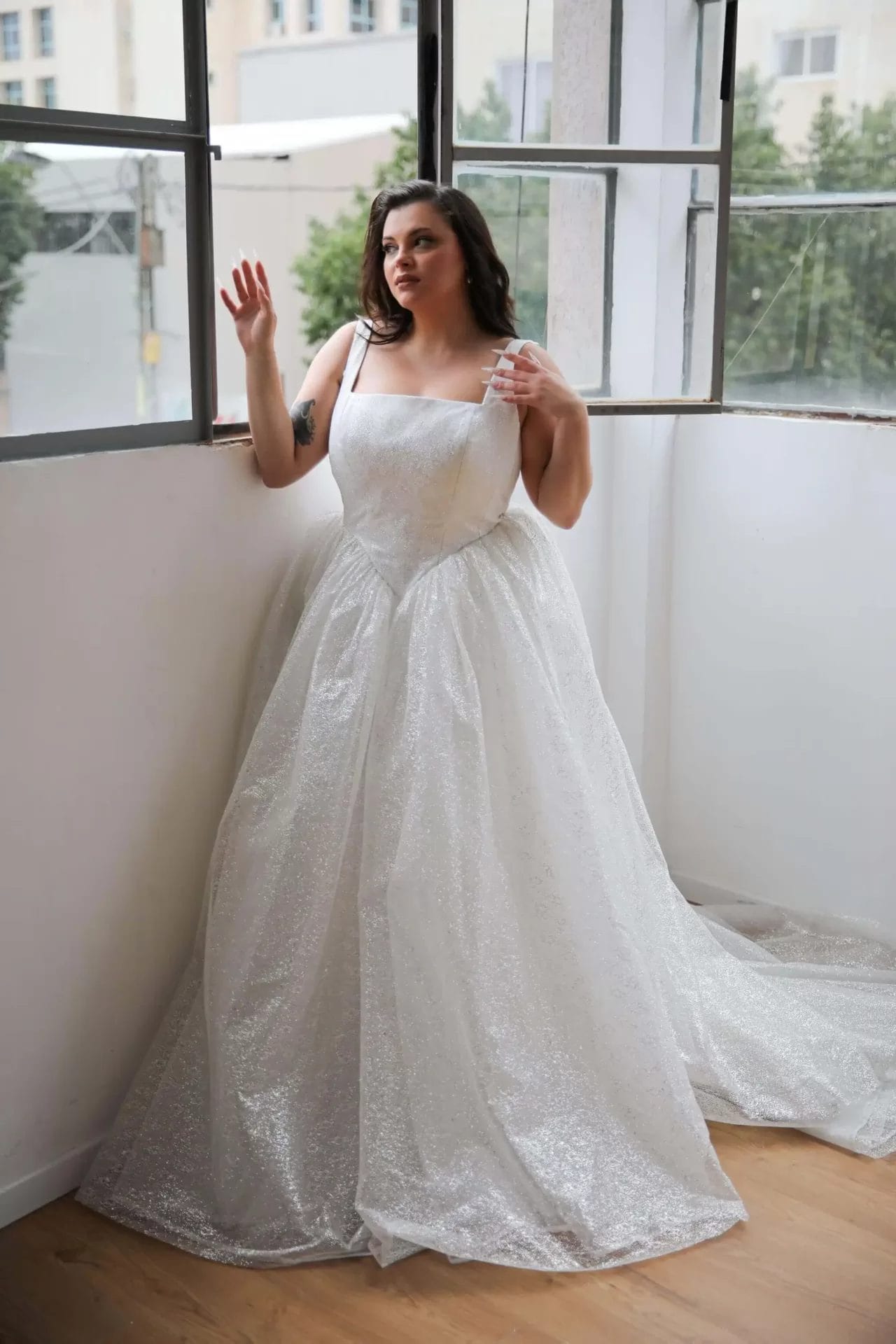 Person wearing a sparkling white gown, standing by a large window, looking outside.