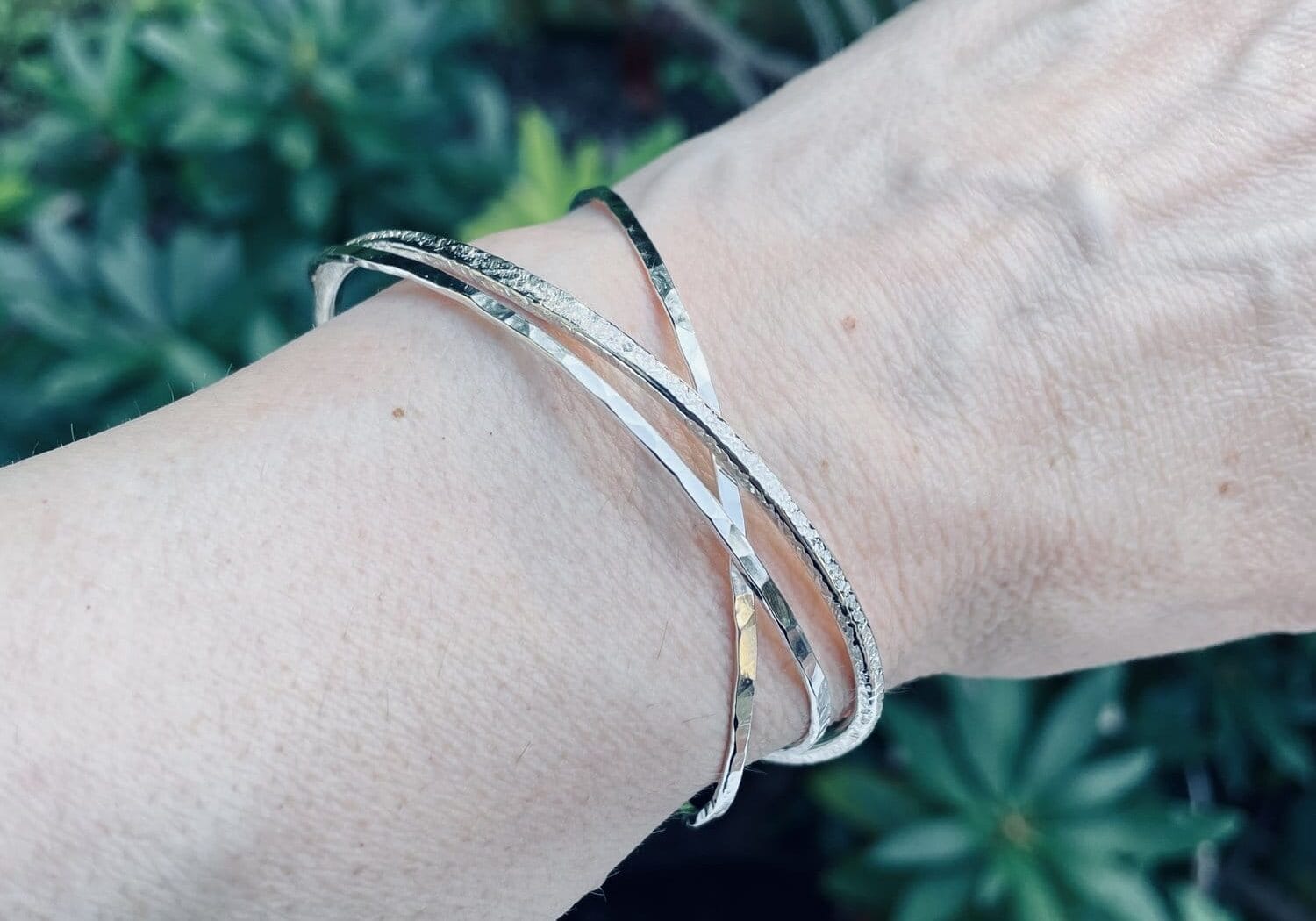 A hand wearing three intertwined silver bangles against a background of green foliage.