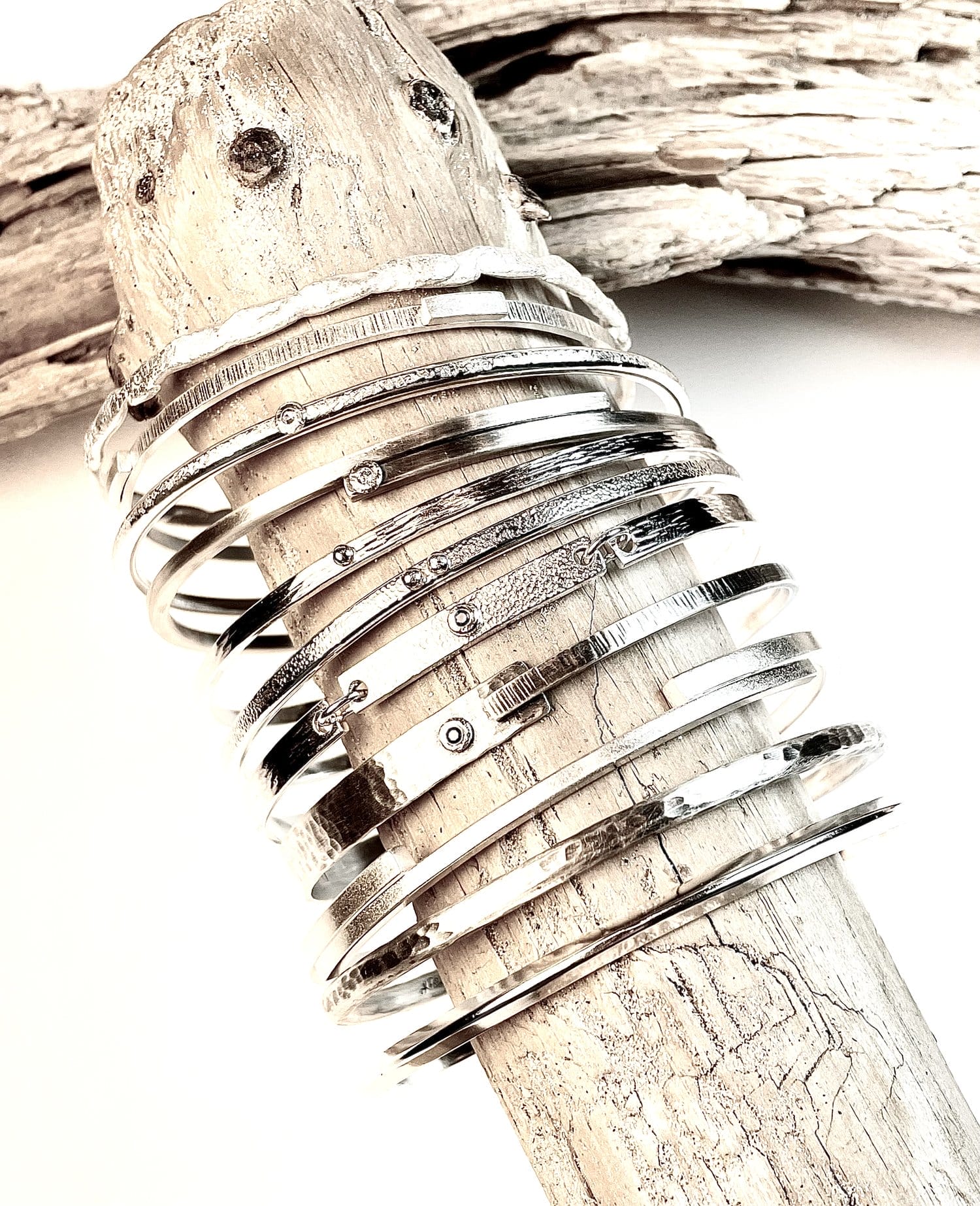 A collection of thin, metallic bangles with various textures and designs is displayed on a piece of driftwood.