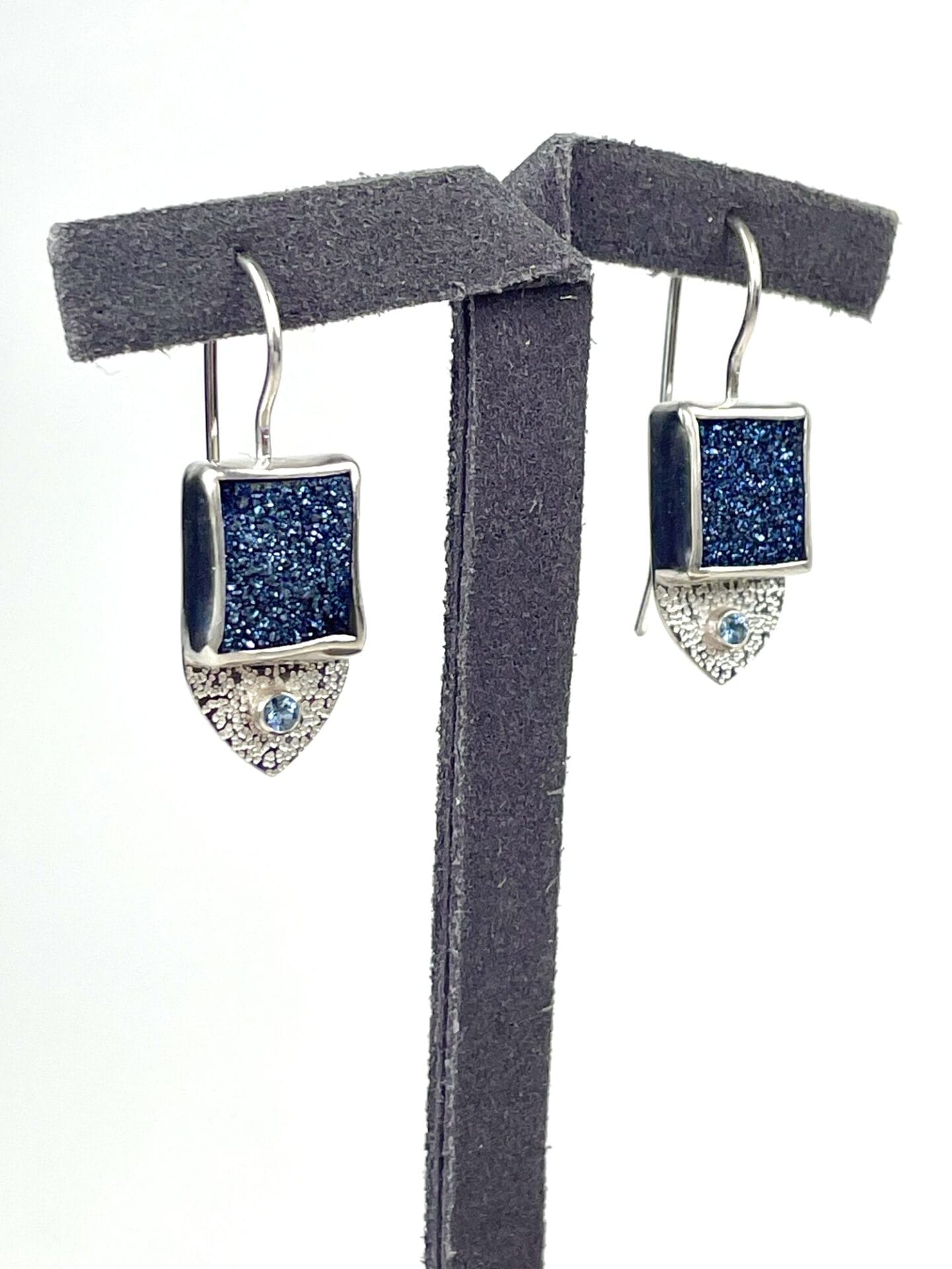 Silver earrings with square, dark blue glittery stones on a black stand. Below each stone is a small light blue gem set in a triangular design.