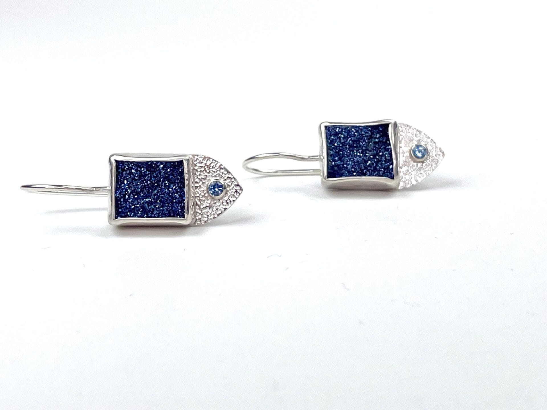 Silver fish-shaped earrings feature rectangular blue glitter centers and small round blue gems for eyes, set against a plain white background.