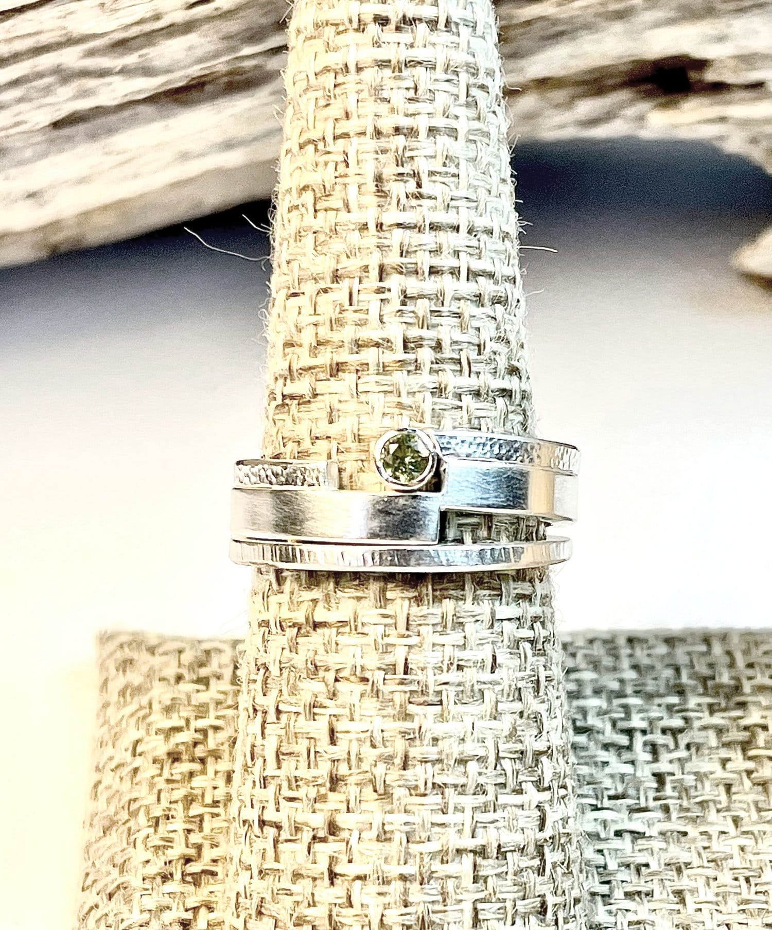 A silver ring with a small green gemstone is displayed on a textured, woven beige stand.