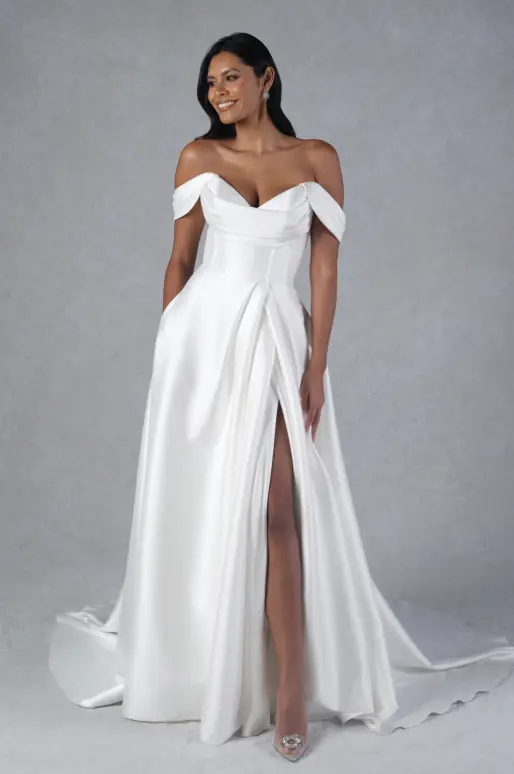 A woman stands in an off-the-shoulder white gown with a thigh-high slit, fitted bodice, and a floor-length skirt, posing against a plain gray background.