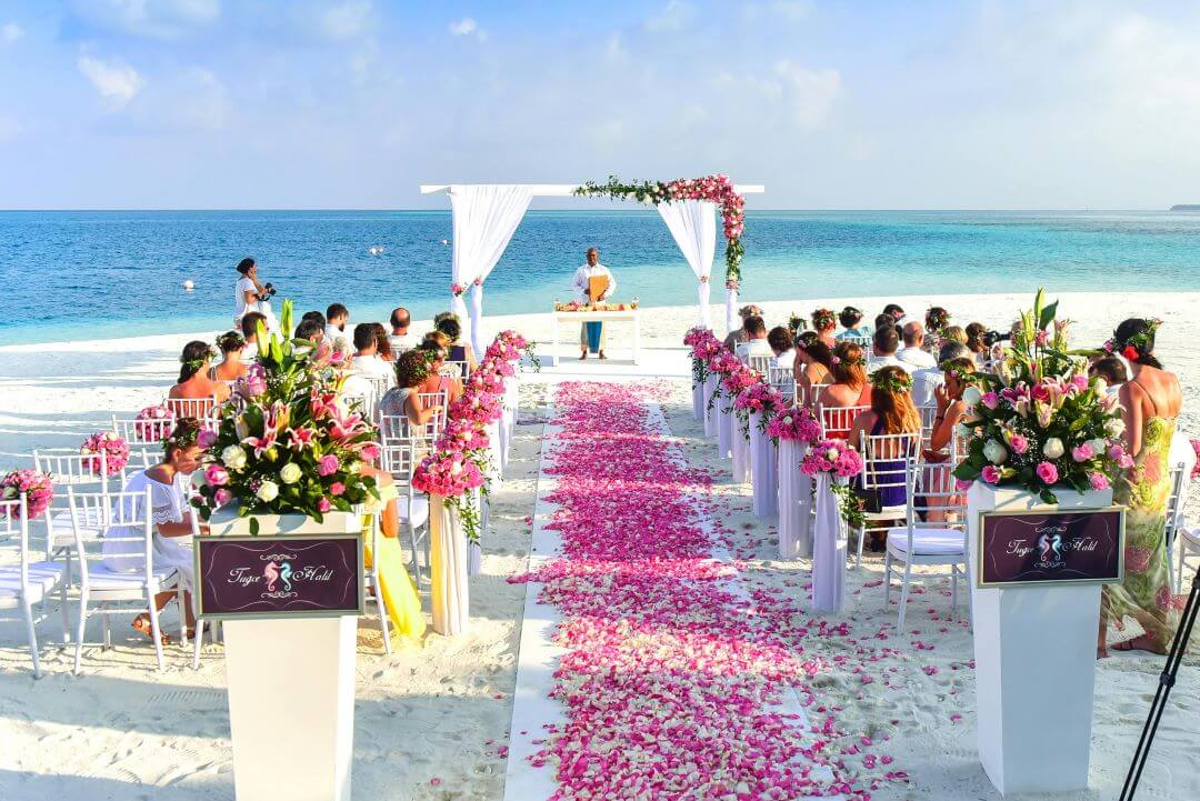 Outdoor beach wedding ceremony setup with chairs, floral arrangements, pink rose petals on the aisle, and guests seated facing an officiant under a white arch by the sea.