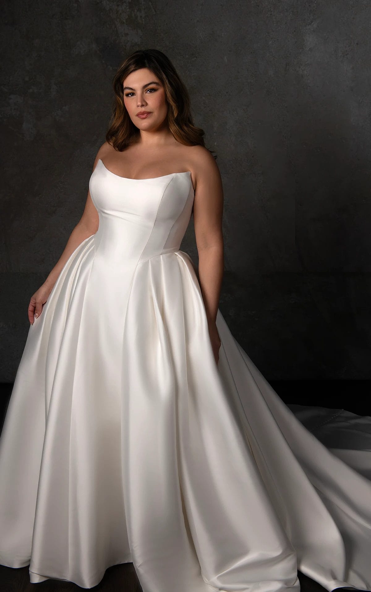 A woman in a strapless, white satin ball gown stands against a dark, textured background.