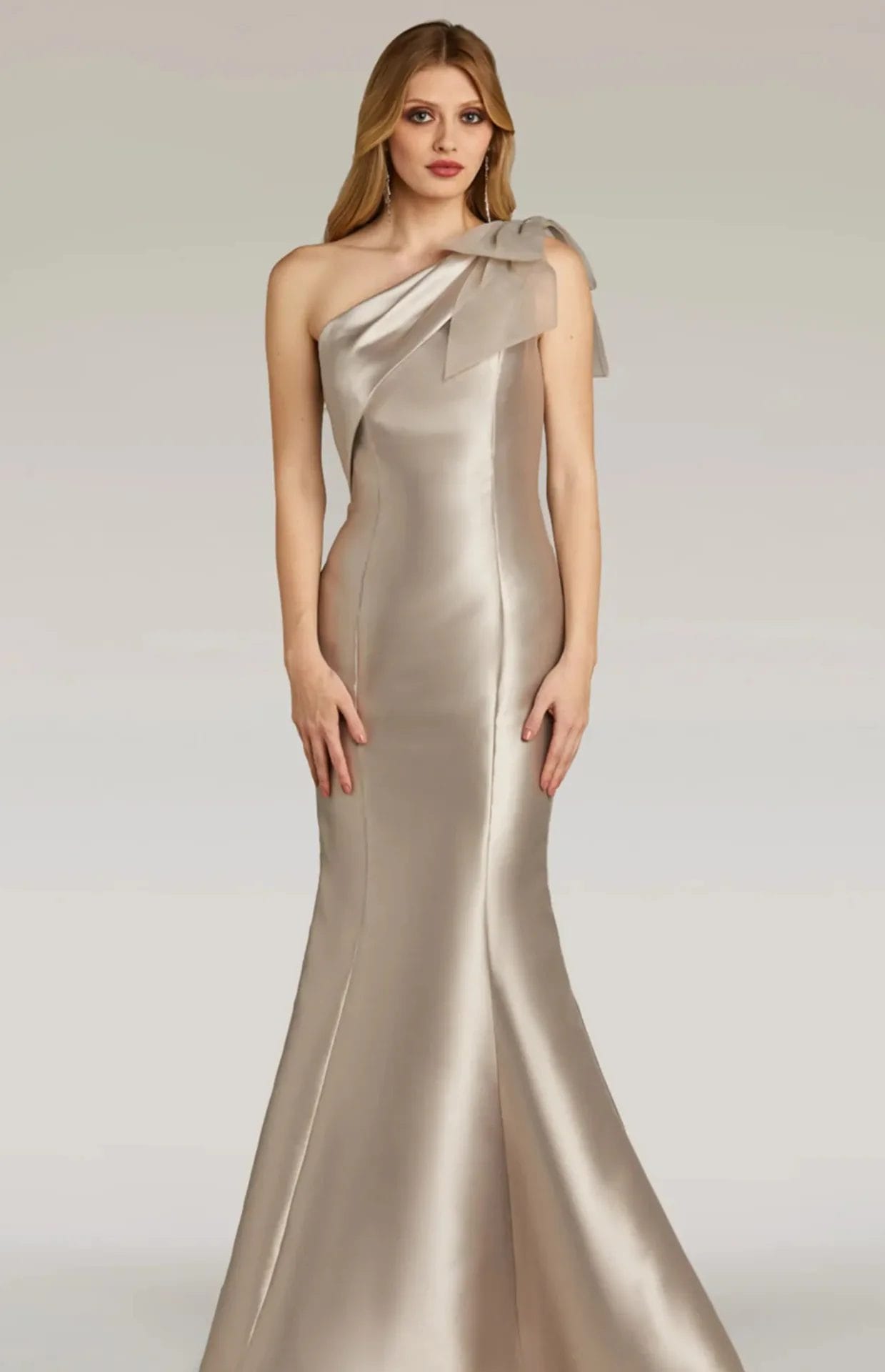 A woman wearing a shiny, champagne-colored, one-shoulder gown with a bow detail at the shoulder stands against a plain background.