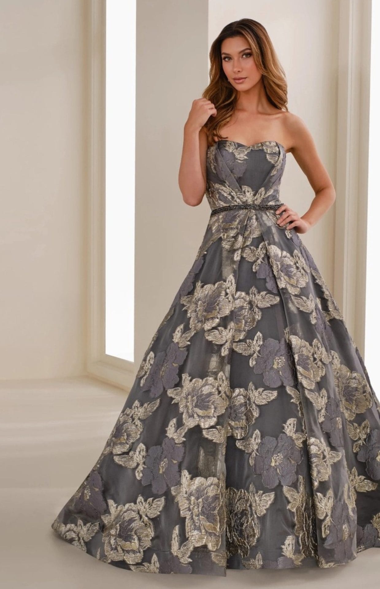 A woman stands indoors wearing a strapless gray ball gown with large metallic gold floral patterns.