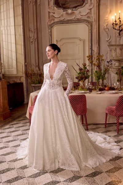 A woman in a long-sleeved, lace wedding gown stands in an ornate room with patterned floors, red chairs, and a decorated dining table—perfect inspiration for destination wedding dress ideas.