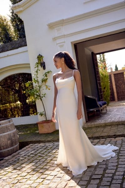 A woman in a simple, elegant white gown stands outdoors on a stone patio beside a building with white walls and an arched doorway, offering stunning inspiration for Destination Wedding Dress Ideas.