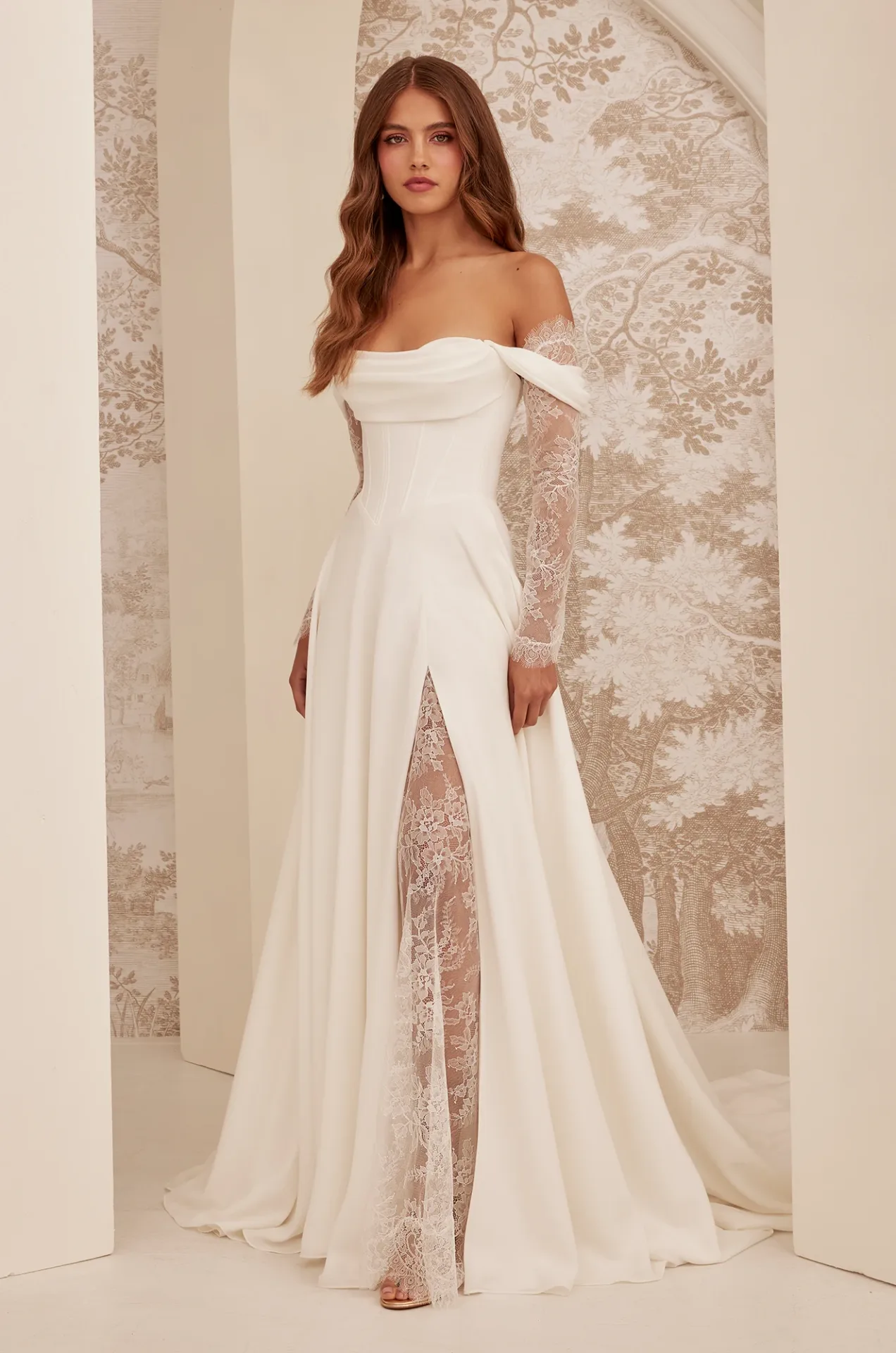 A woman wearing an off-shoulder white gown with lace sleeves and a front slit stands indoors against a decorative, neutral-toned background.