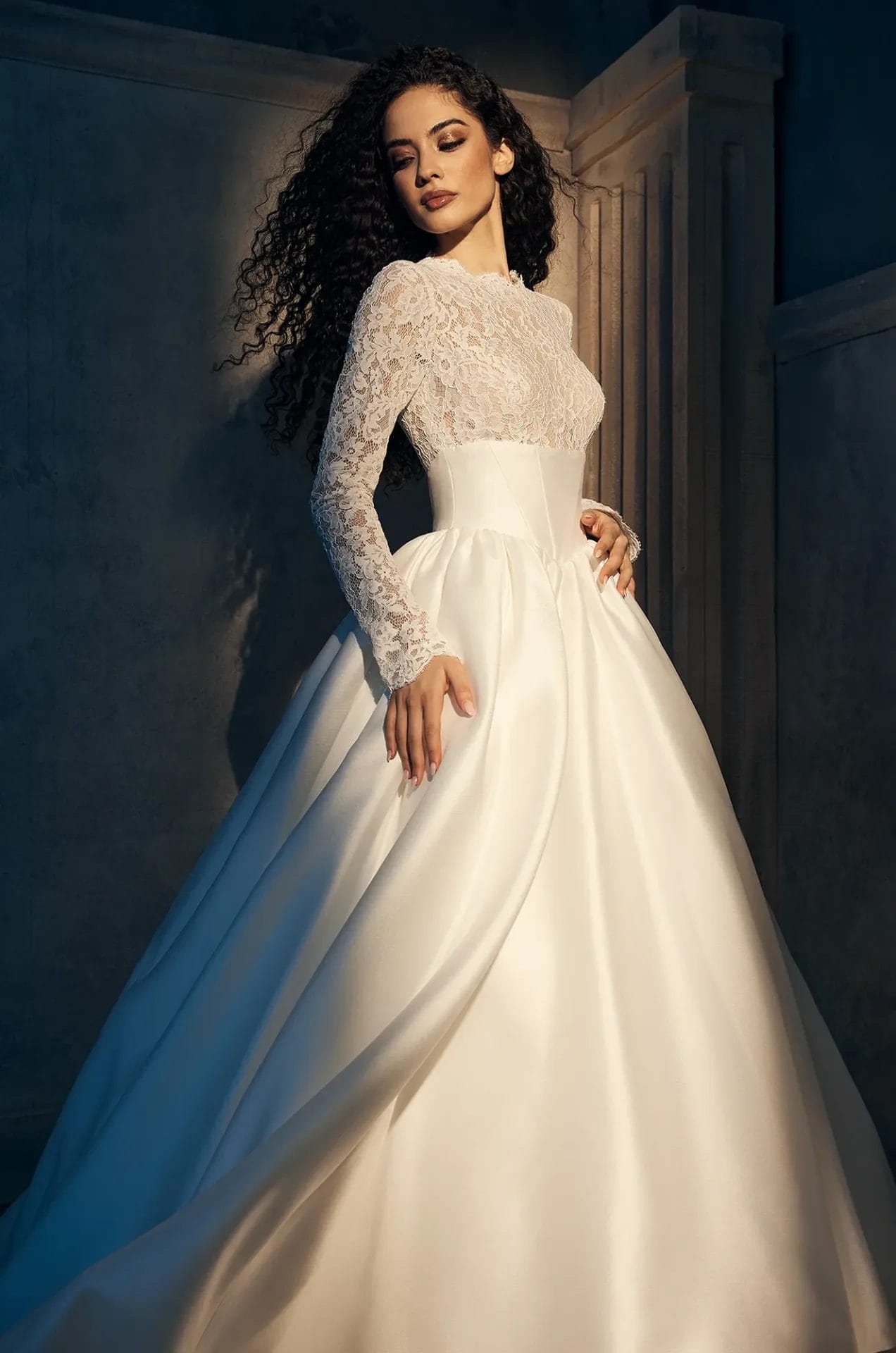A woman in a long-sleeve lace and satin ball gown wedding dress stands in a dimly lit, elegant setting with architectural details in the background.
