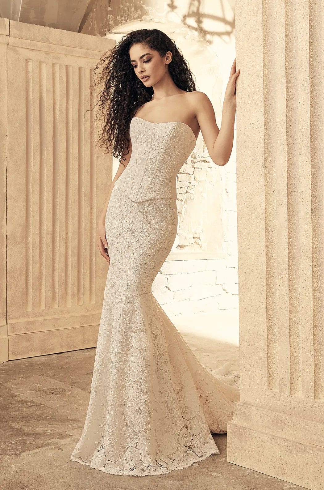 A woman with long curly hair wears a strapless, form-fitting lace wedding gown and stands against a stone column in a softly lit setting.