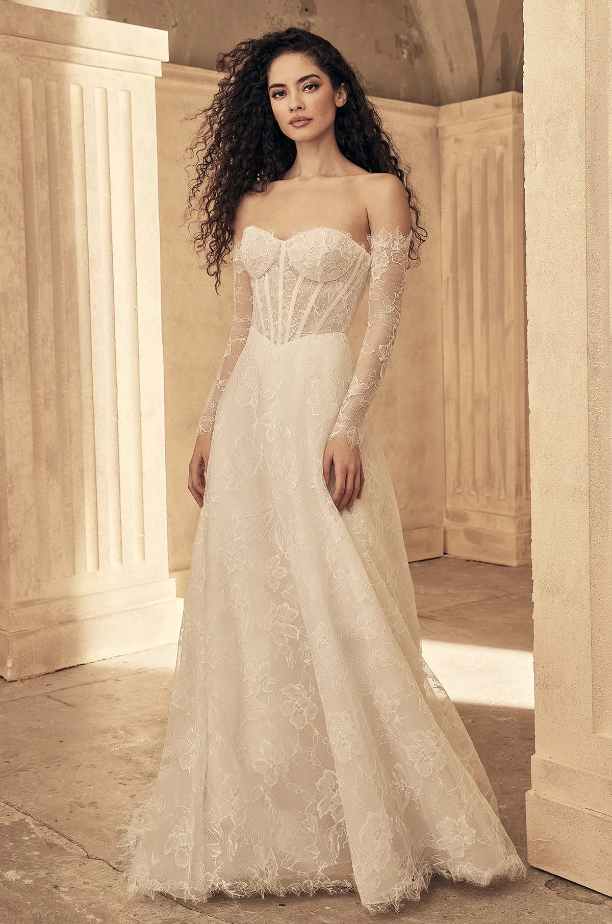 A woman wearing an off-the-shoulder, long-sleeve lace wedding dress with a fitted bodice and flowing skirt stands in a light-colored, columned room.
