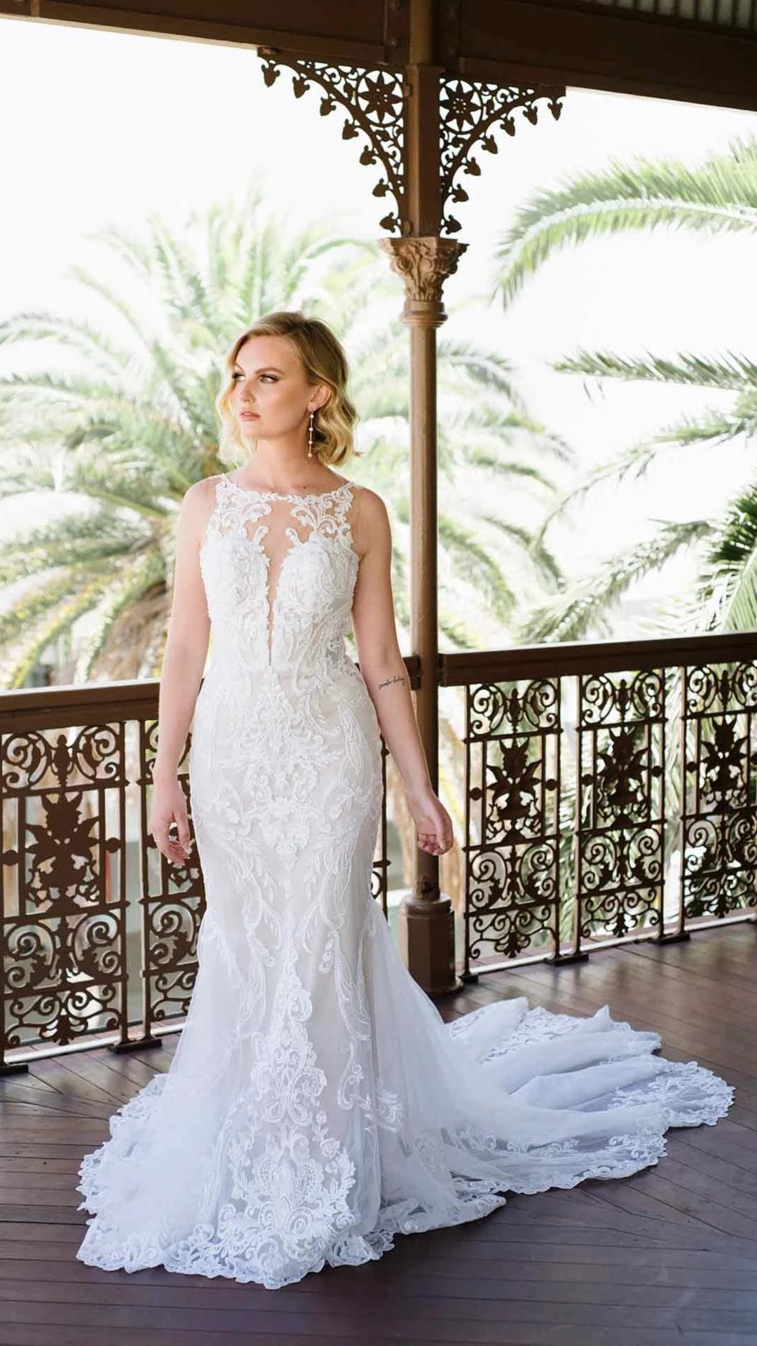 A woman in a fitted, sleeveless lace wedding dress with a long train stands on a wrought-iron balcony with palm trees in the background.