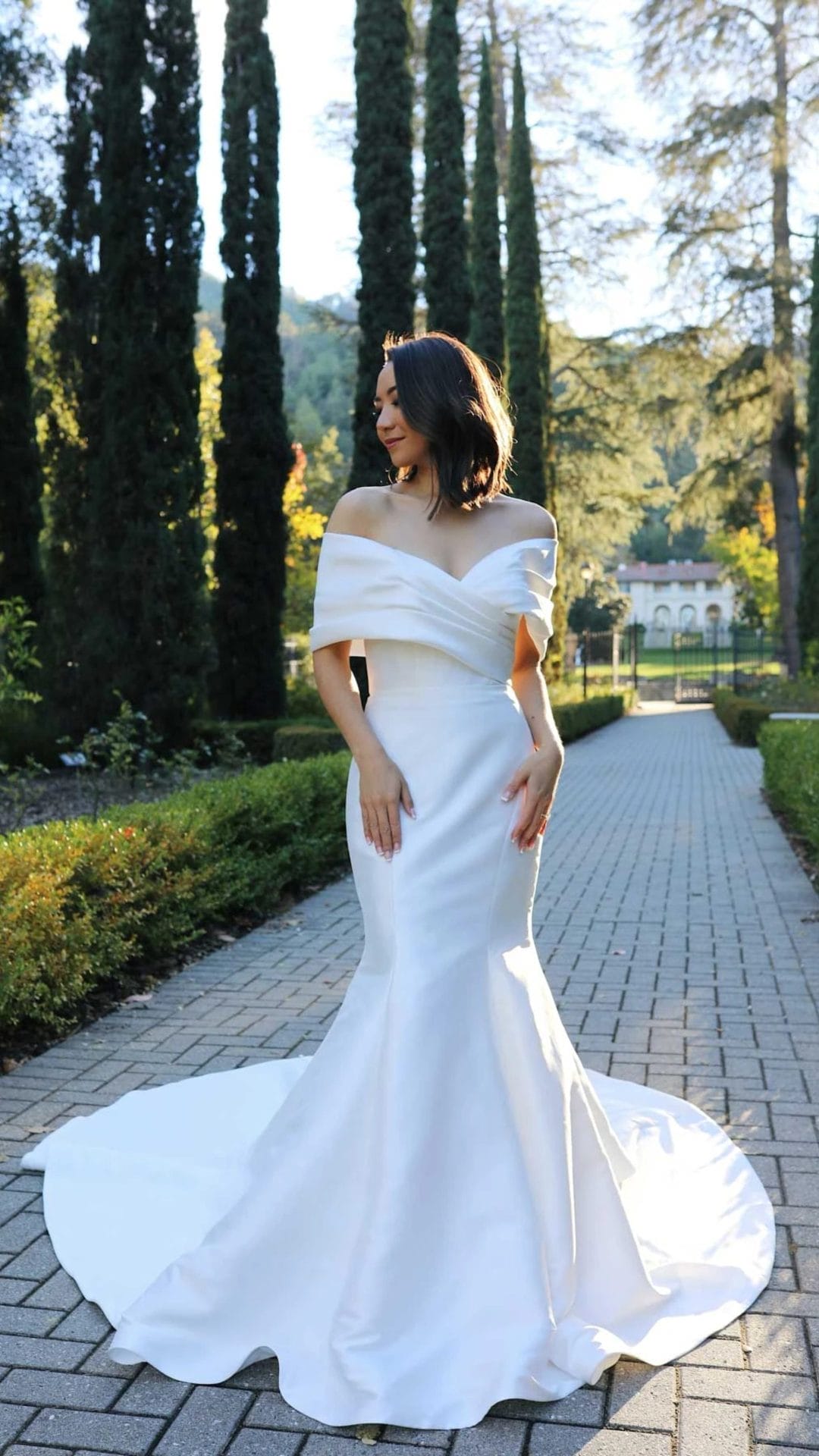A woman in an off-shoulder white wedding gown stands on a brick pathway in a garden with tall trees and a building in the background.