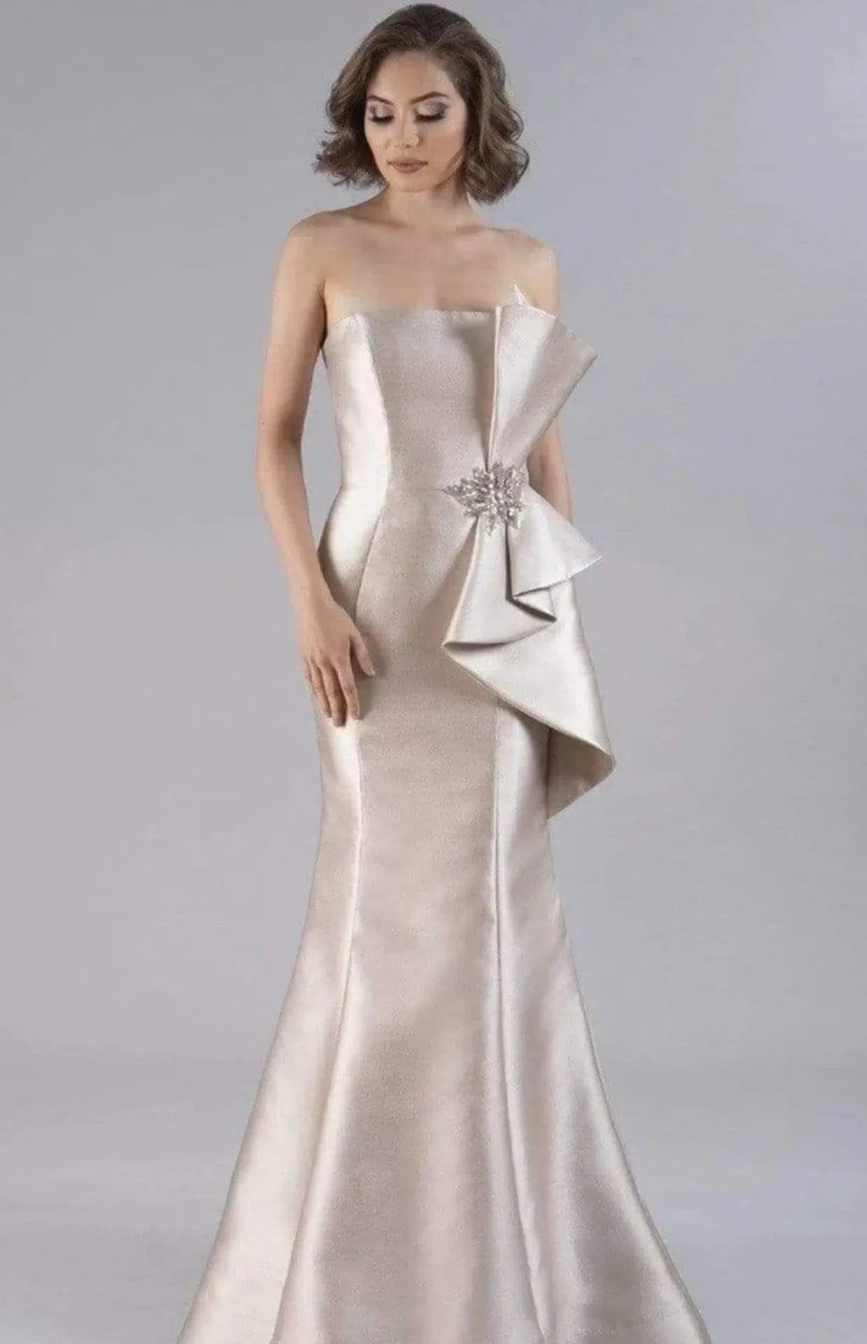 Woman wearing a strapless, floor-length, metallic gown with a large side bow and jeweled brooch detail, standing against a plain gray background.
