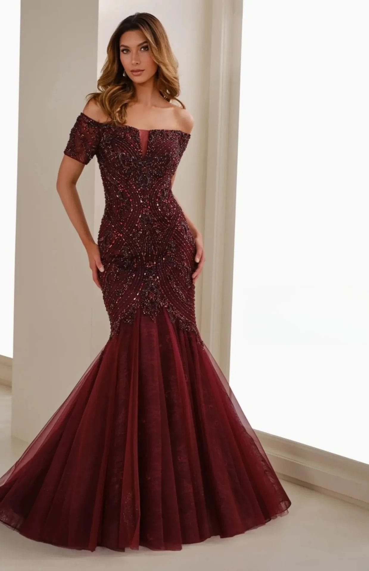 A woman stands indoors wearing an off-the-shoulder, burgundy mermaid gown with beaded embellishments and a tulle skirt.