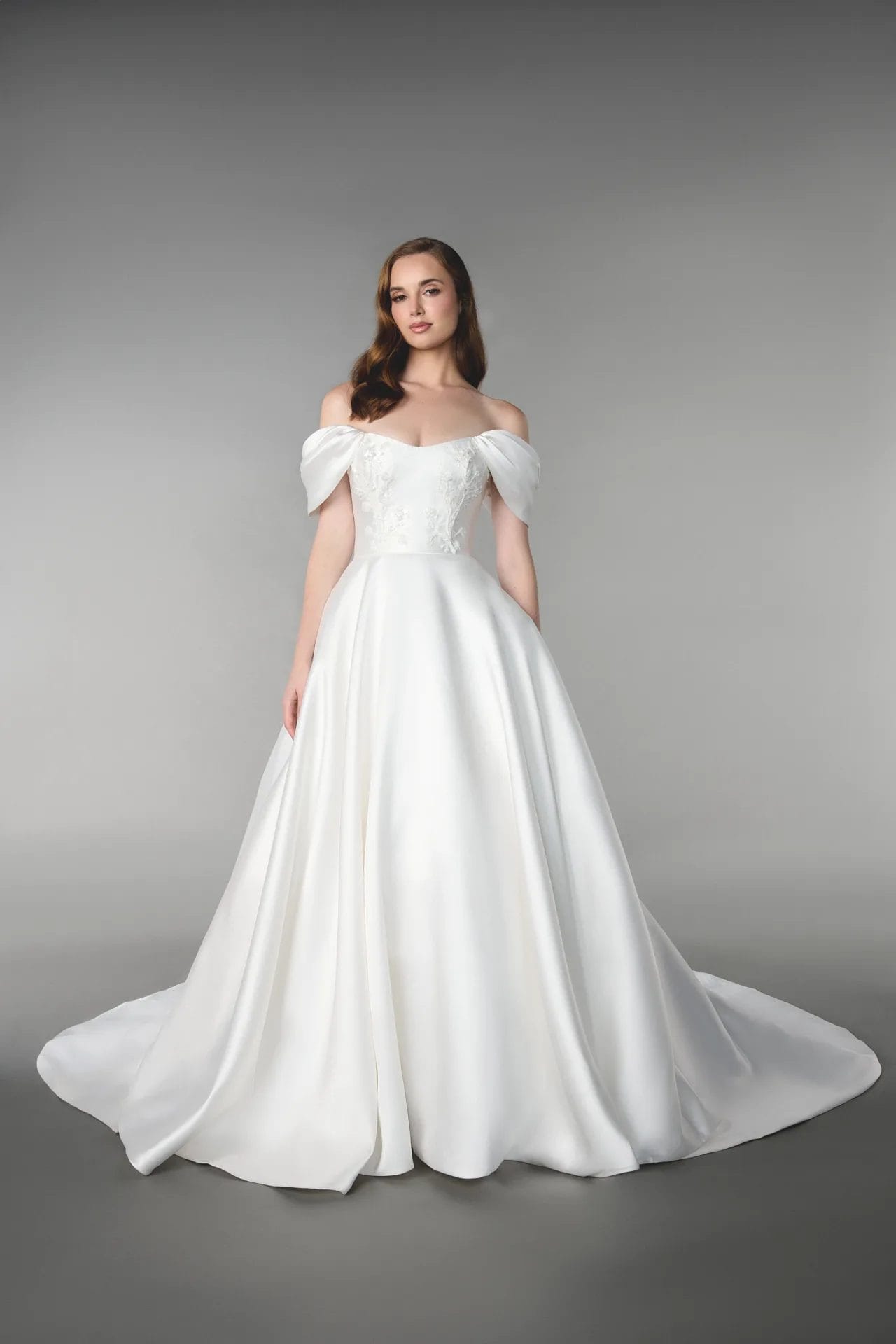 A woman stands wearing an off-the-shoulder white wedding gown with a fitted bodice, embroidered details, and a full skirt with a train against a plain gray background.