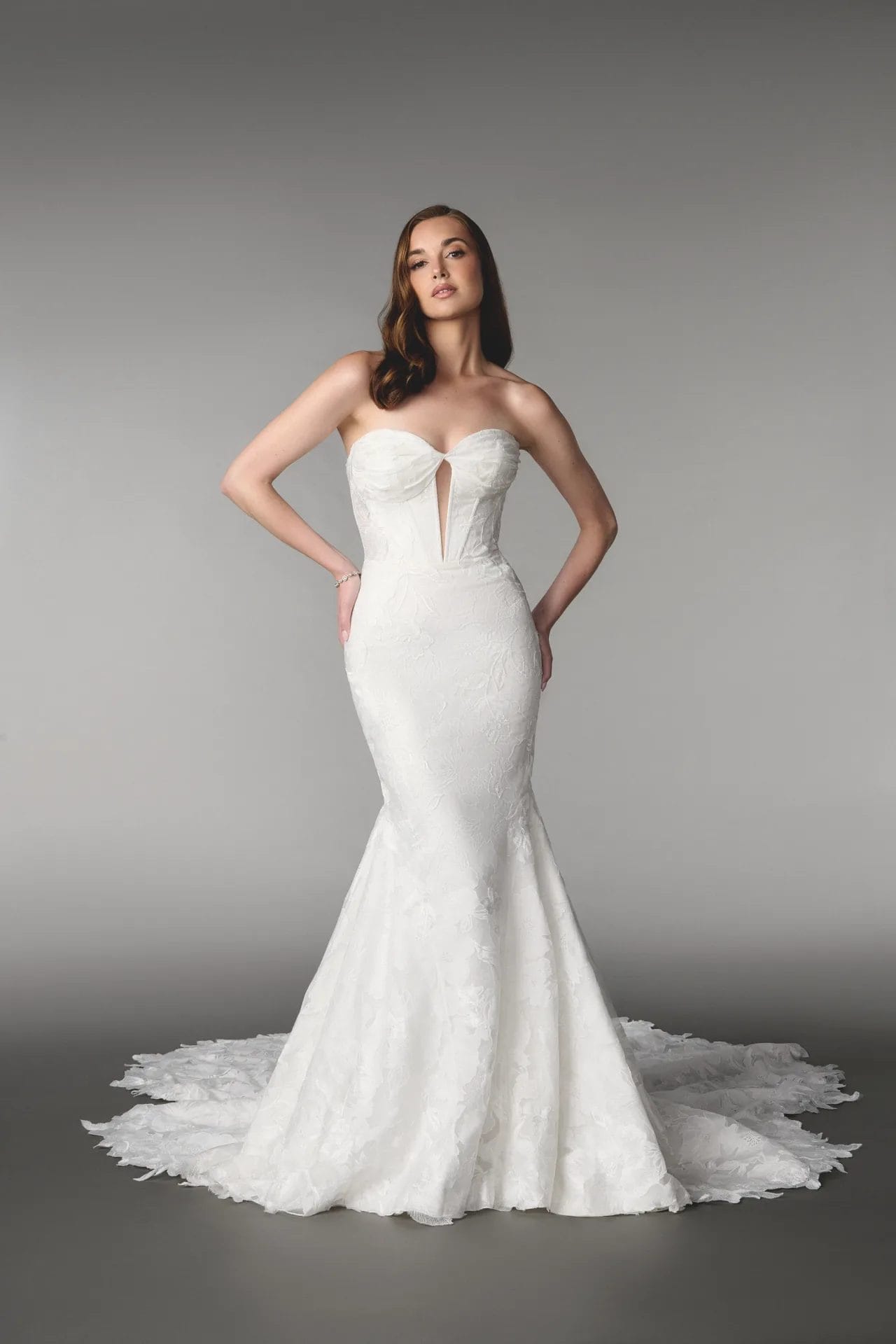 A woman wearing a strapless, form-fitting white wedding dress with a front slit and a long lace train stands against a plain gray background.