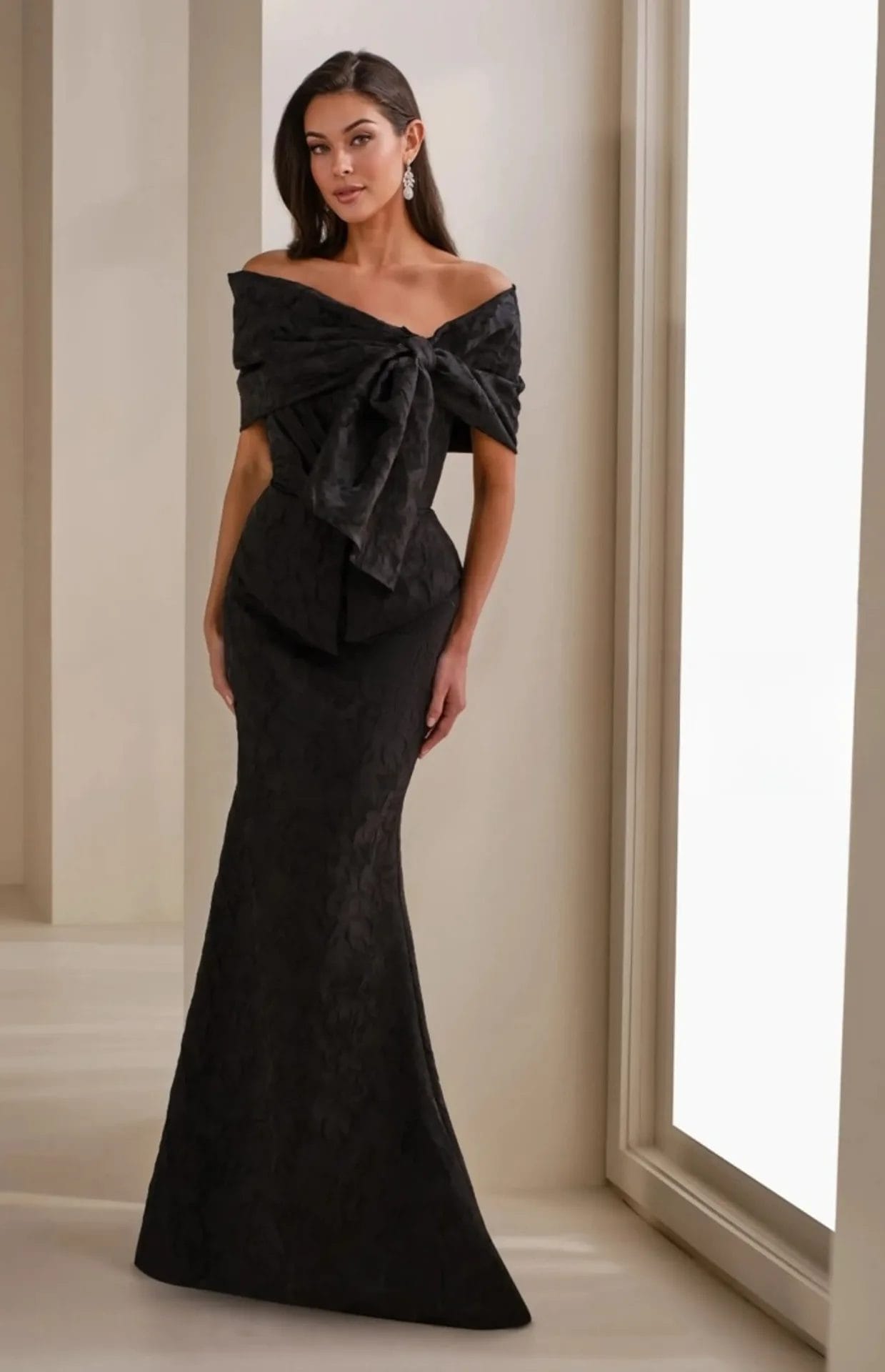 A woman stands indoors wearing an off-the-shoulder, floor-length black gown with a large bow detail on the bodice.
