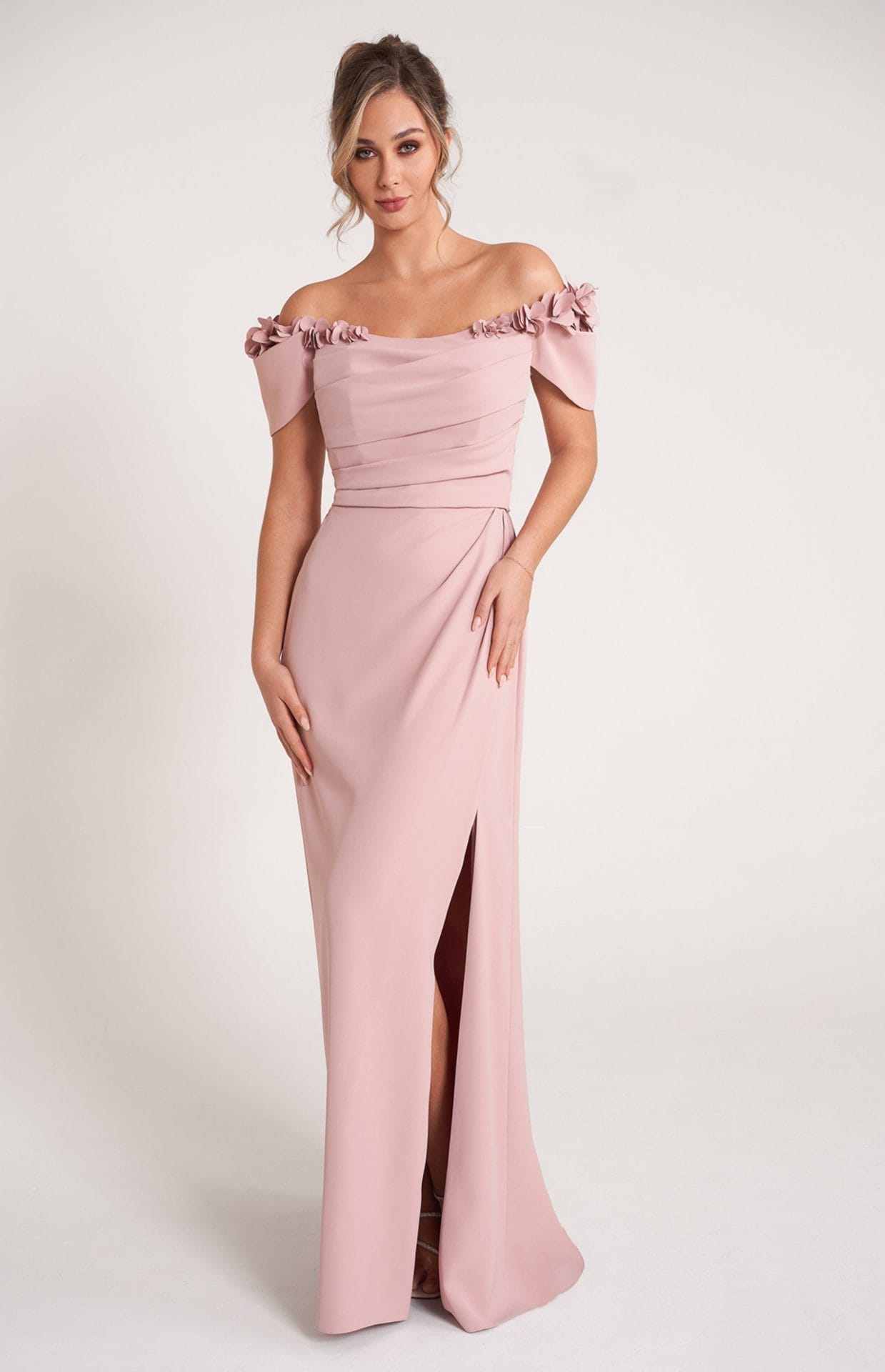 A woman wearing a blush pink off-the-shoulder gown with floral details on the sleeves and a front slit, standing against a plain white background.