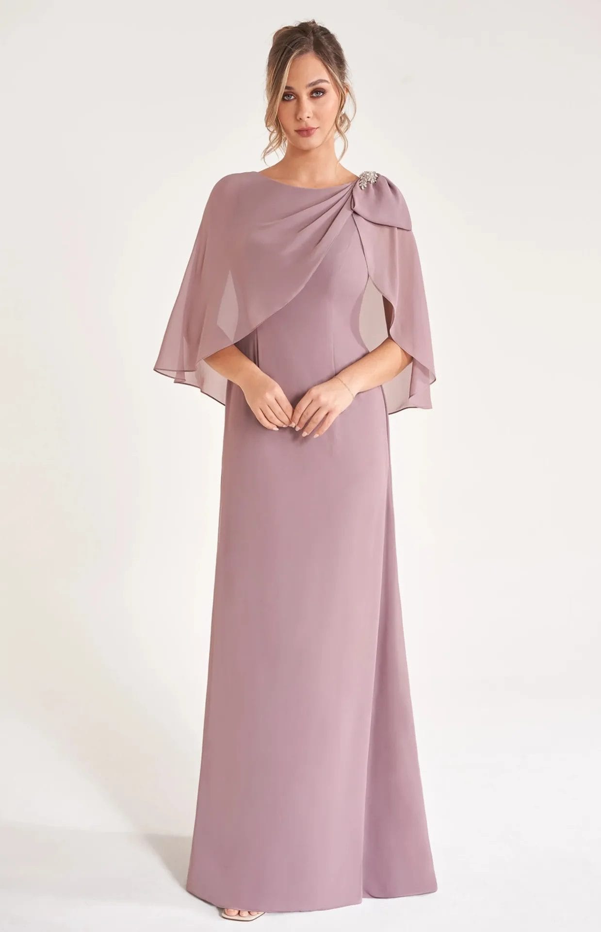 A woman stands wearing a floor-length mauve gown with a sheer capelet overlay, adorned with a jeweled brooch on one shoulder, against a plain light background.