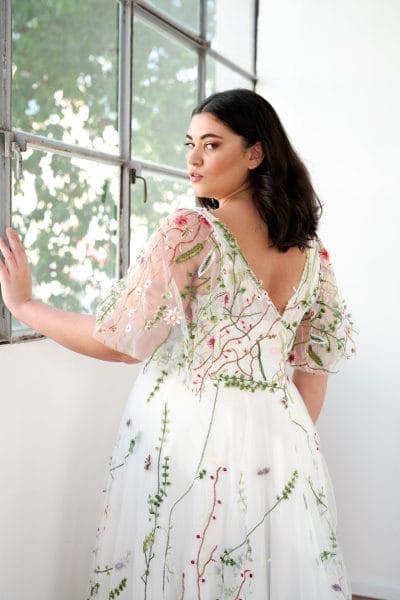 Woman in a white dress with floral embroidery stands by a window, sunlight filtering through the glass and greenery outside—capturing effortless elegance and inspiring Destination Wedding Dress Ideas.